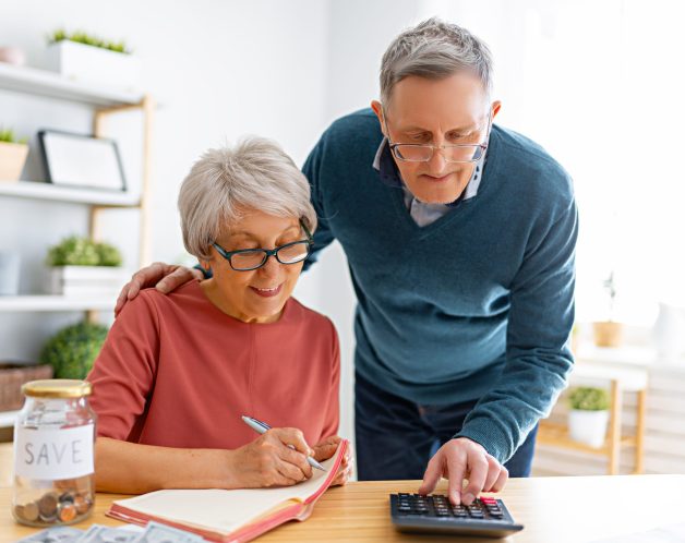 Elderly married couple sitting at the desk with a paper receipt in hands are calculating expenses, managing the family budget.